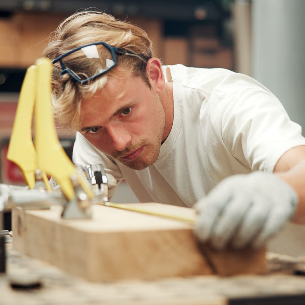 Person mit Schutzbrille misst ein Holzstück in einer Werkstatt.